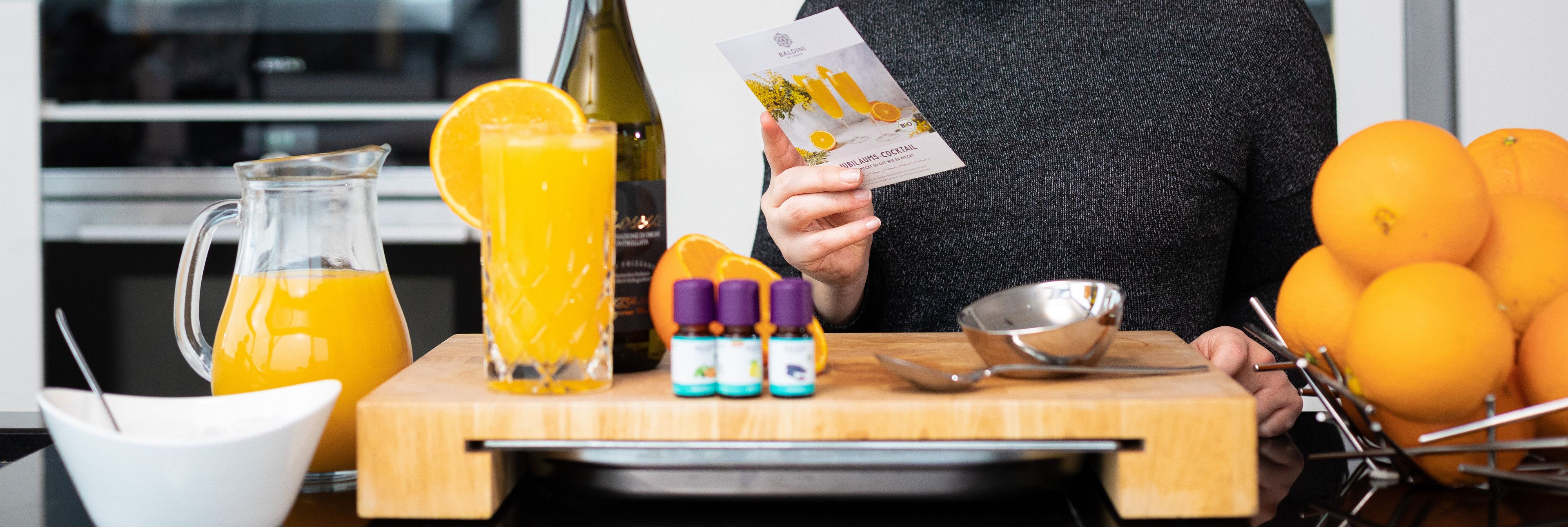 Person bereitet Orangensaft-Getränk zu, hält Duftkarte, mit frischen Orangen, Saftkrug und ätherischen Ölen auf Holzbrett.