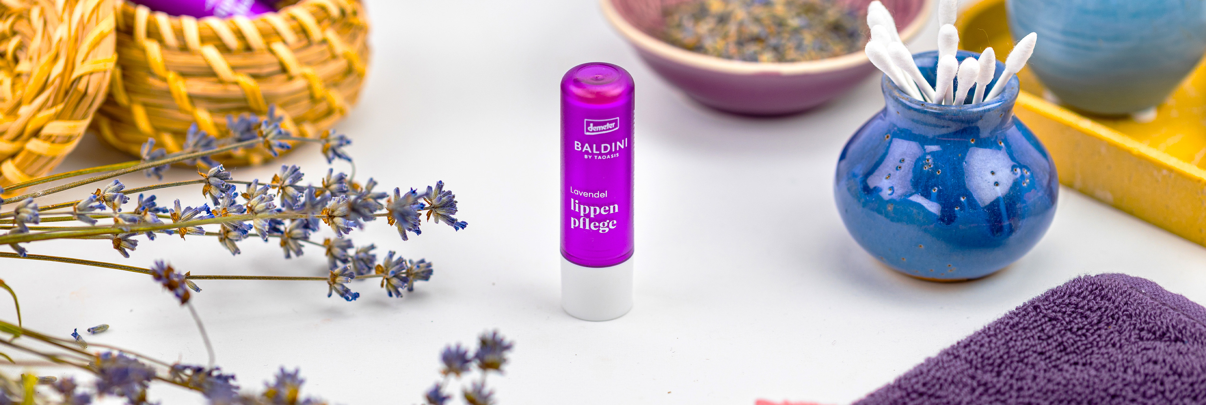 Baldini Lippenpflegestift Lavendel mit Lavendelblüten und Kosmetikutensilien.