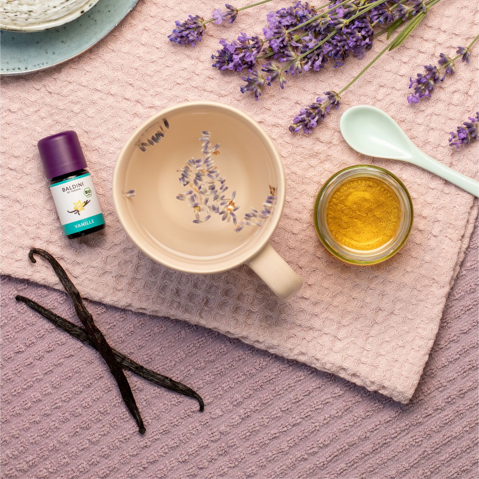 Bio-Essenz Vanille und eine Tasse Tee. Lavendelblüten liegen auf dem Tisch.