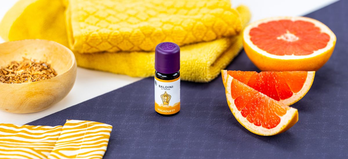 Baldini Yogaduft ätherisches Öl auf einer dunklen Yogamatte, umgeben von Grapefruithälften, einem Holzschälchen mit getrockneten Pflanzenteilen und gelben Handtüchern – frische, natürliche Atmosphäre für Yoga und Entspannung.