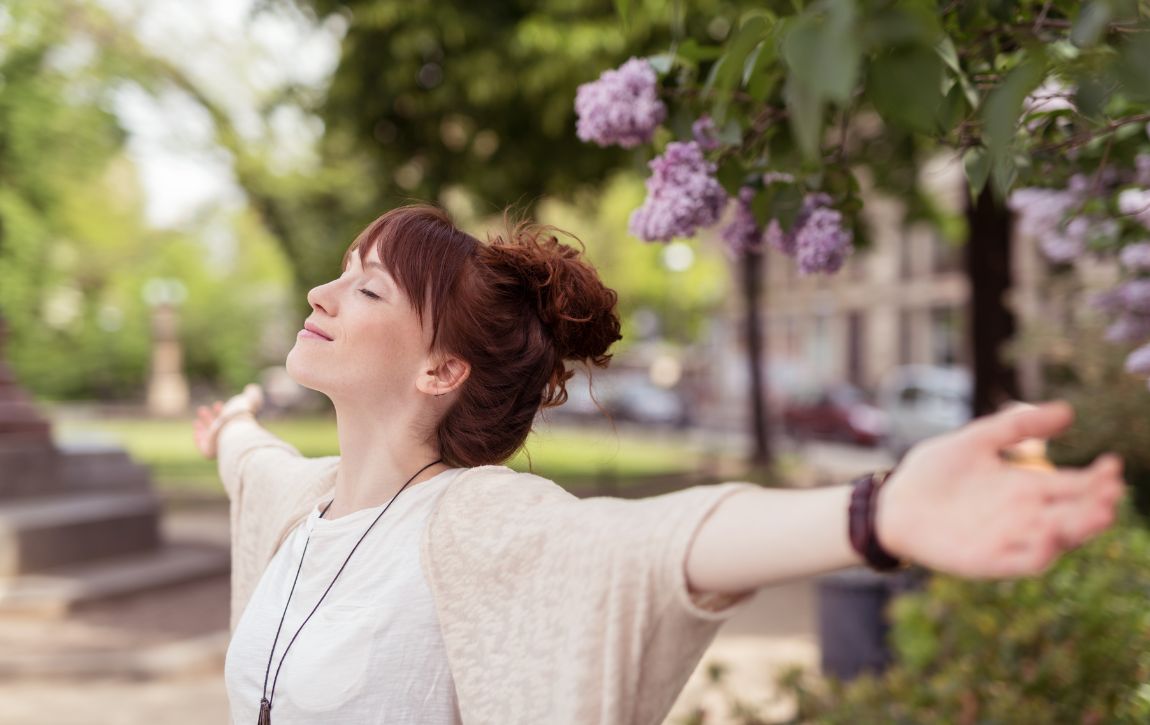 Frau mit ausgestreckten Armen, geschlossenen Augen atmet tief ein.  Natur ist zu sehen.