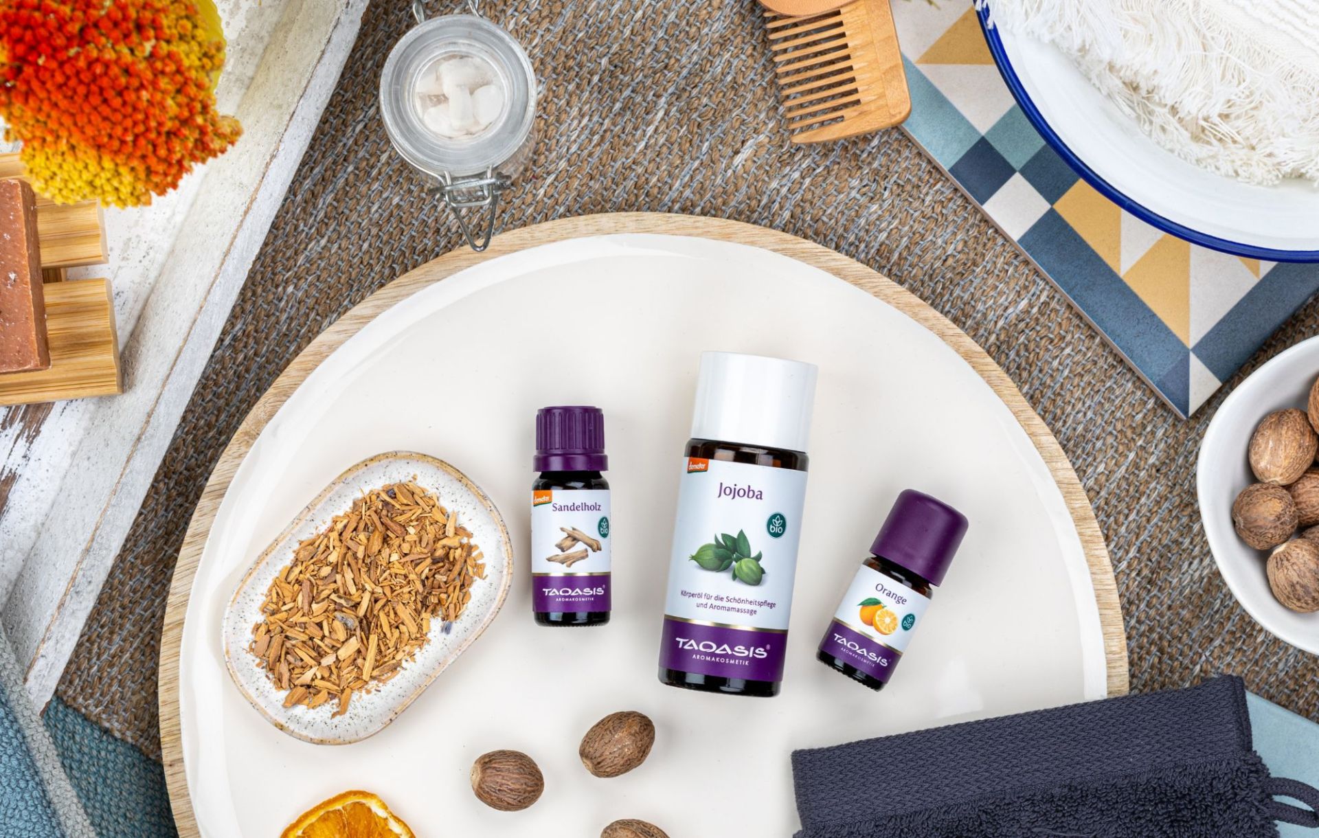AEO Sandelholz, Basisöl Jojoba und AEO Orange auf einem weißen Teller. Umgeben von Orangenschreiben, Nüssen und Badezimmerutensilien