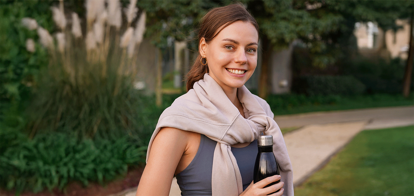 Lächelnde Frau in Sportkleidung steht mit Trinkflasche in einem grünen Park.