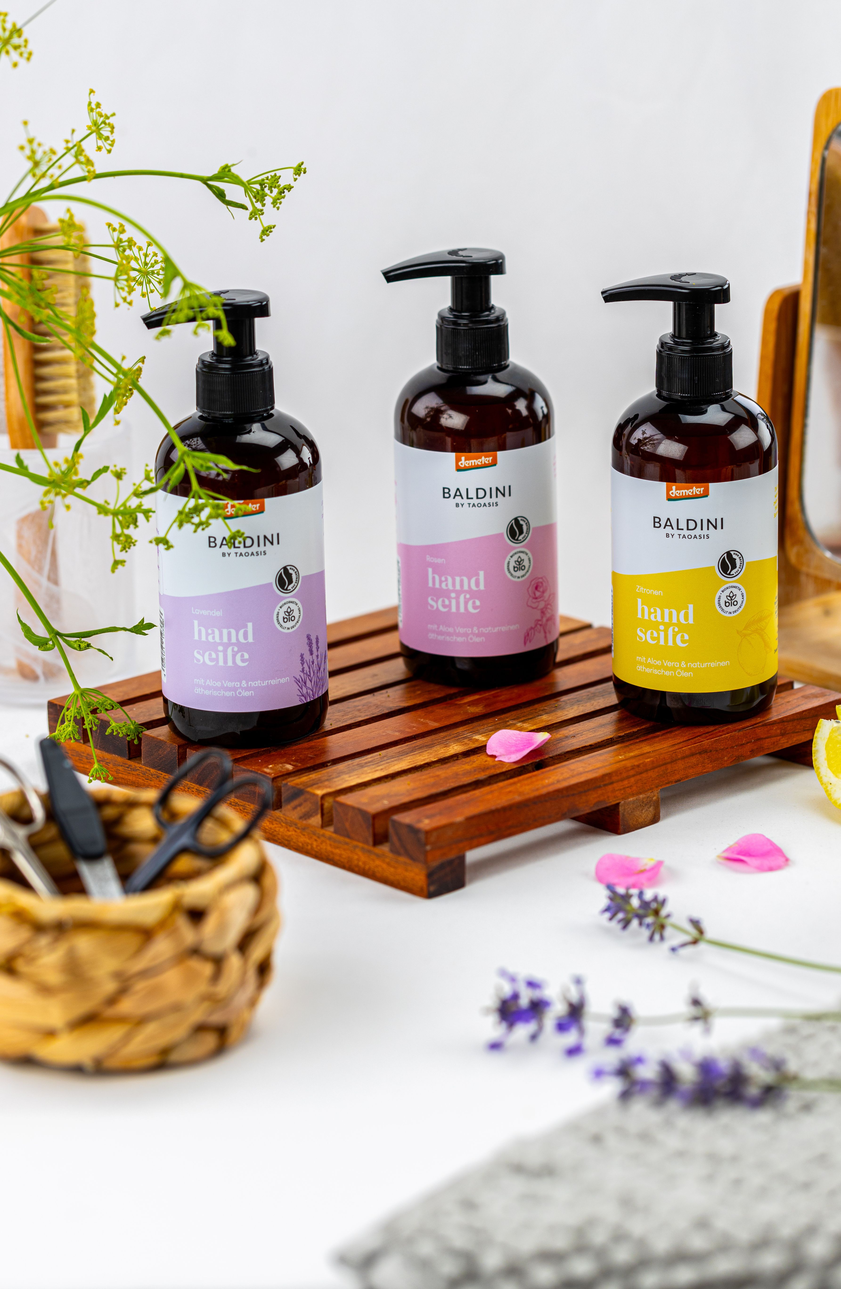 Drei Flaschen Baldini Handseife (Rose, Zitrus, Lavendel) auf einem Holztablett, umgeben von Blumen und einem Korb mit Kosmetikutensilien.