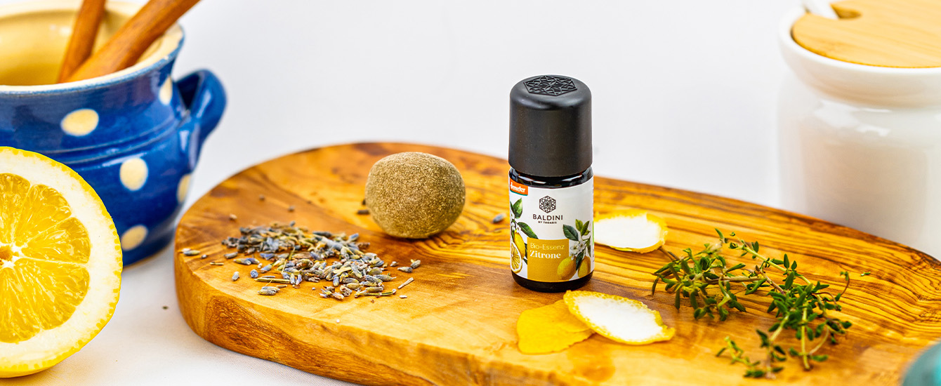 Fläschchen mit Baldini Bio-Essenz Zitrone auf einem Holzbrett, umgeben von Zitrone, Lavendel und frischen Kräutern.