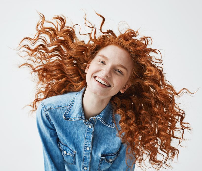 Rothaarige Frau, in Jeans Jacke mit Locken die glücklich in die Kamera schaut.