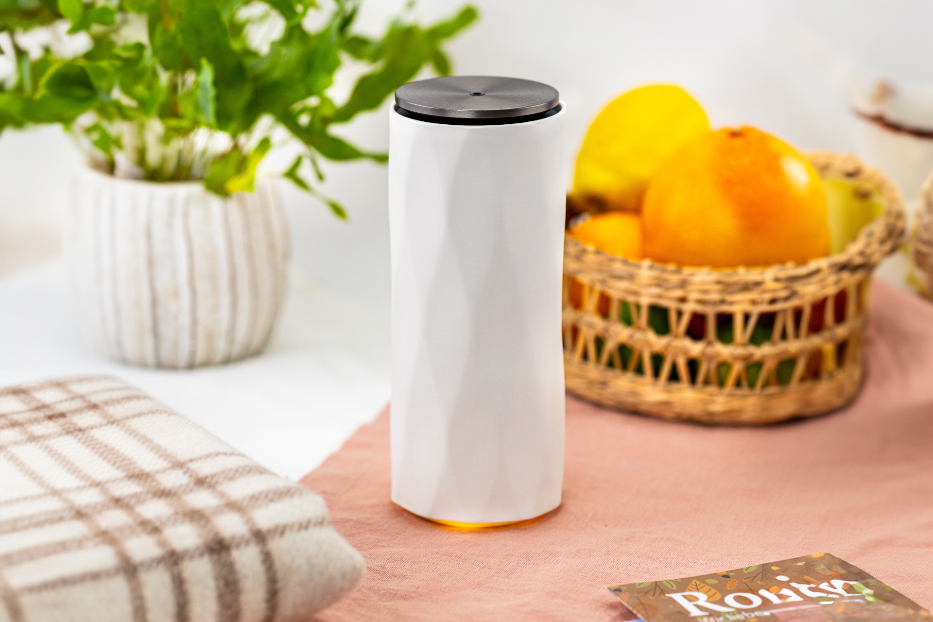 Baldini AirFriend PURE Aroma-Diffusor in modernem, weißem Design mit dunklem Deckel, stilvoll in einem gemütlichen Ambiente platziert – mit Obstkorb, Decke und Zimmerpflanze im Hintergrund.