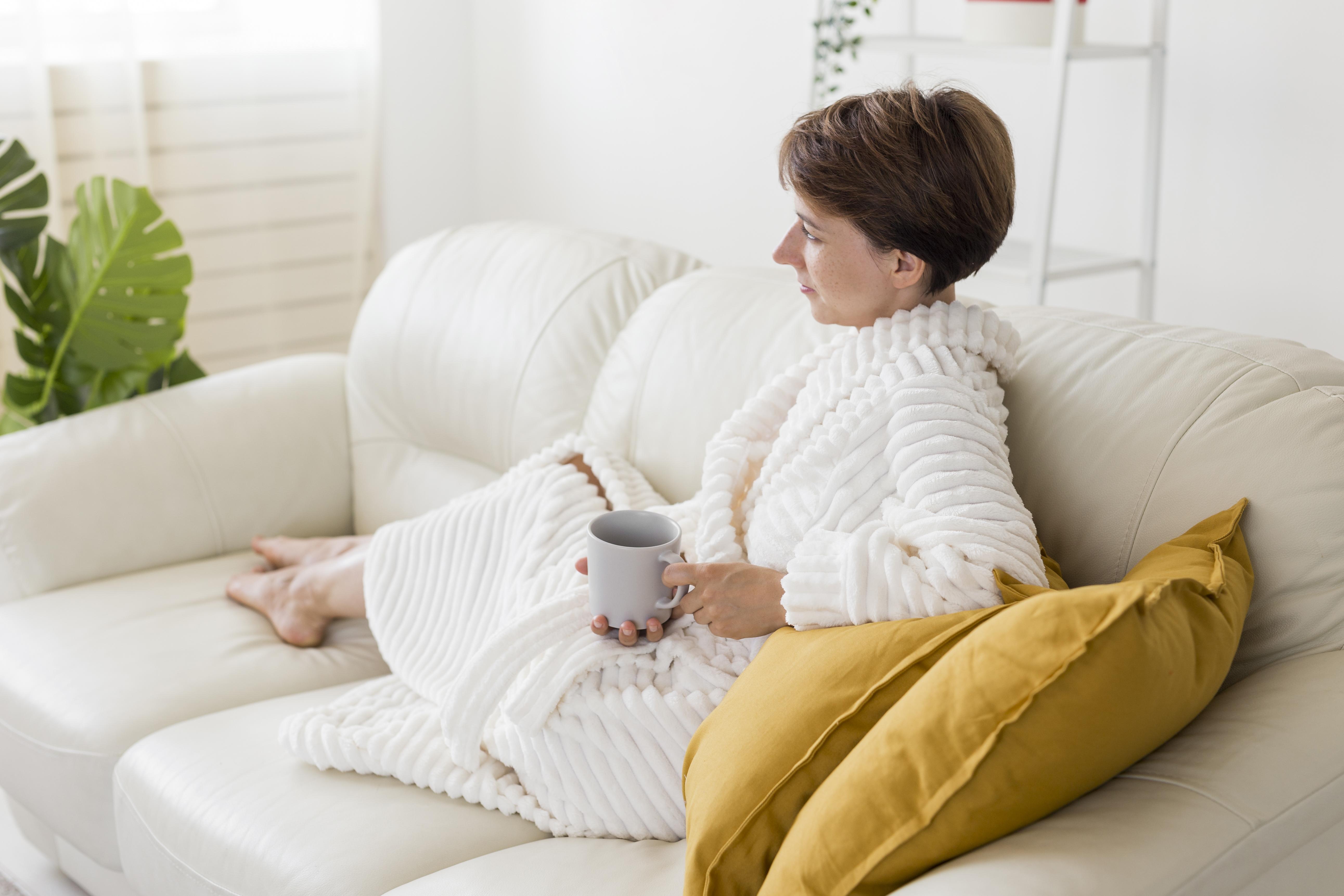 Dame sitzt mit Bademantel und einer Tasse in der Hand auf dem Sofa.