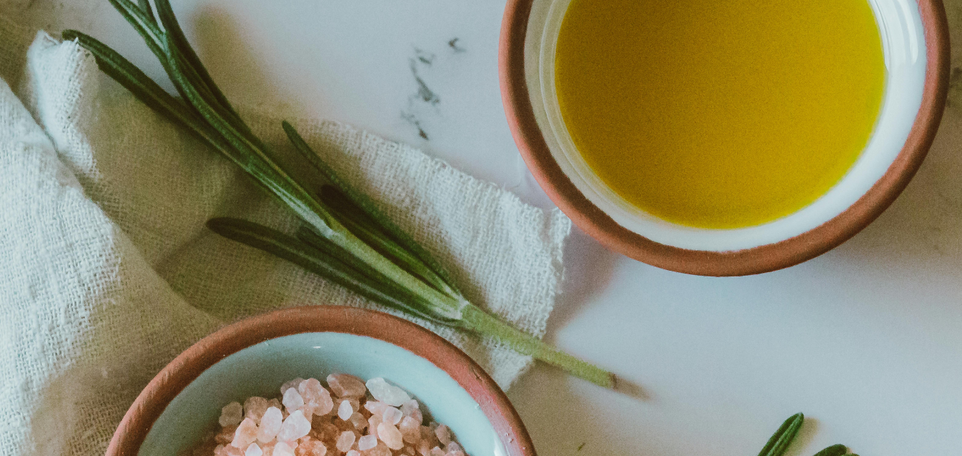 Schale mit goldenem Öl, Rosmarinzweig und Meersalz auf hellem Hintergrund.