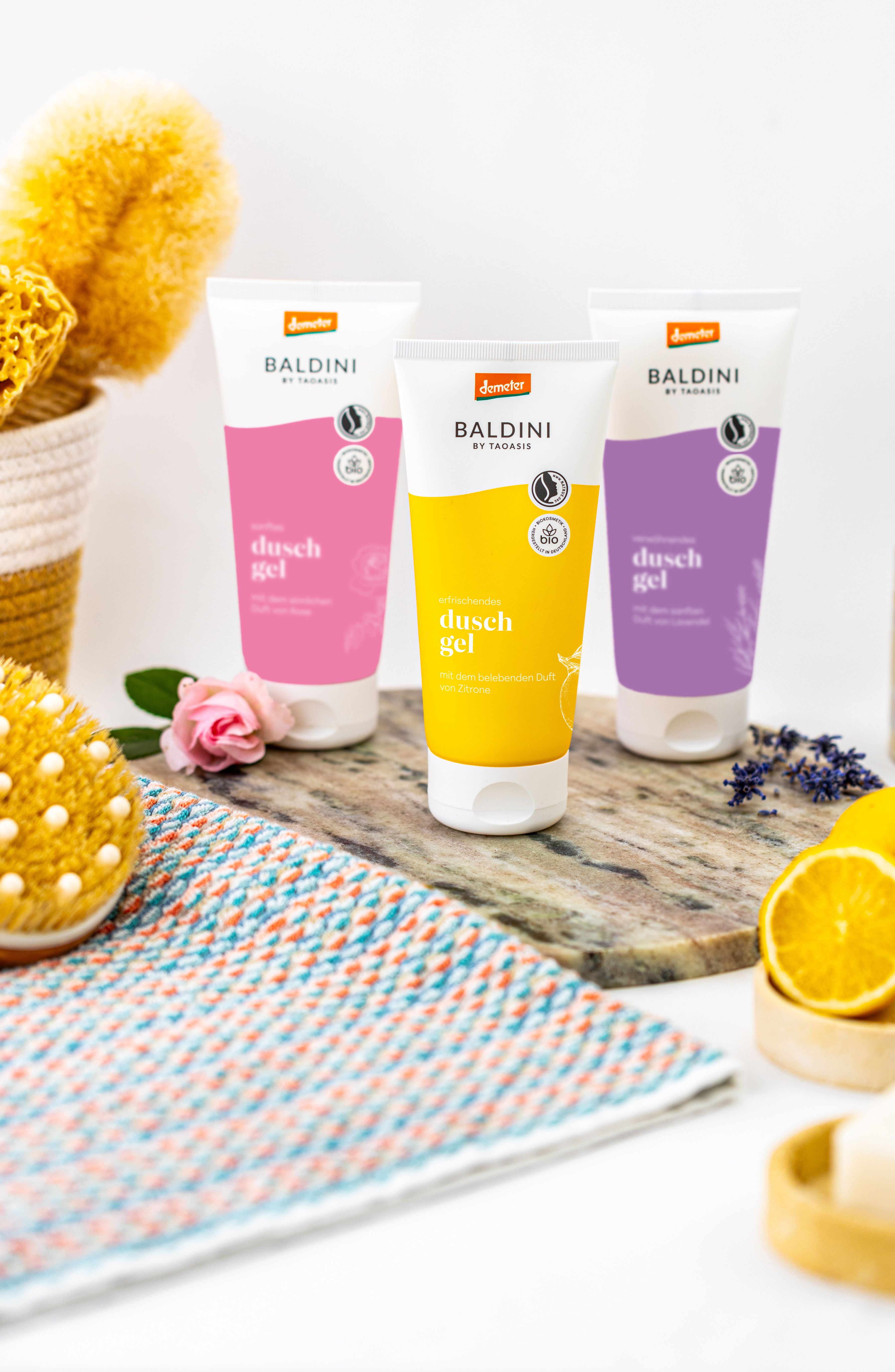 Badlini Duschgel (Rose, Lavendel, Zitrus) auf einem Holztablett. Im Hintergrund ist eine Blume zu sehen uns im Vordergrund liegt ein Handtuch.