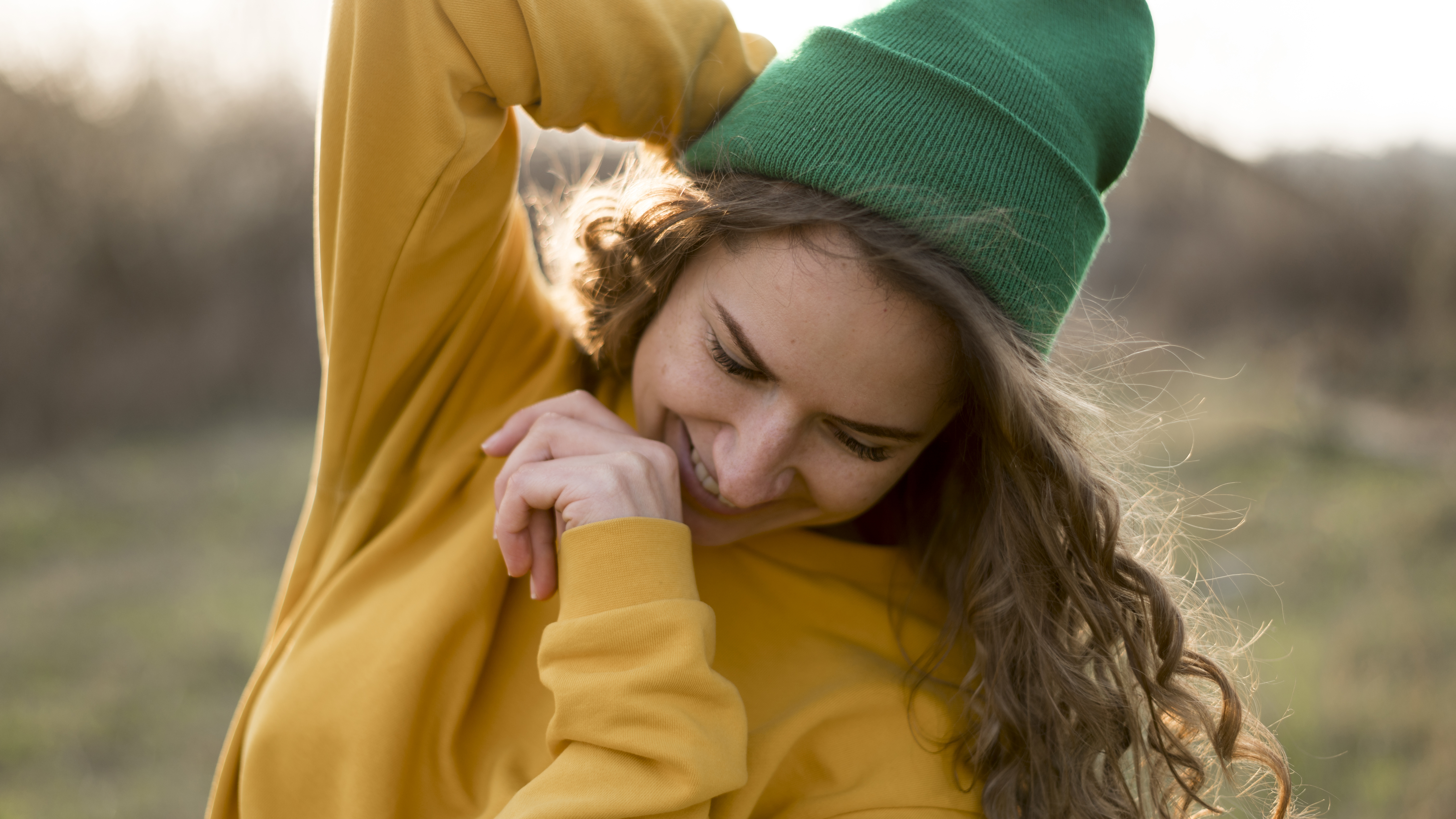 Lächelnde Frau mit grünem Beanie und gelbem Pullover steht draußen in der Natur und bewegt fröhlich ihre Arme.
