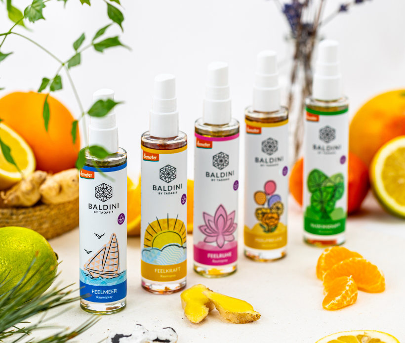 Fünf Baldini Raumsprays von TAOASIS mit frischen Früchten und Pflanzen dekoriert.