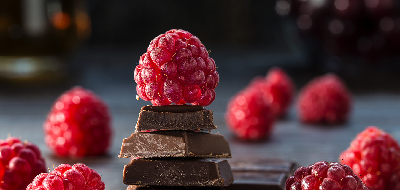 Frische Himbeere auf gestapelten Schokoladenstücken, umgeben von weiteren Himbeeren vor dunklem Hintergrund.