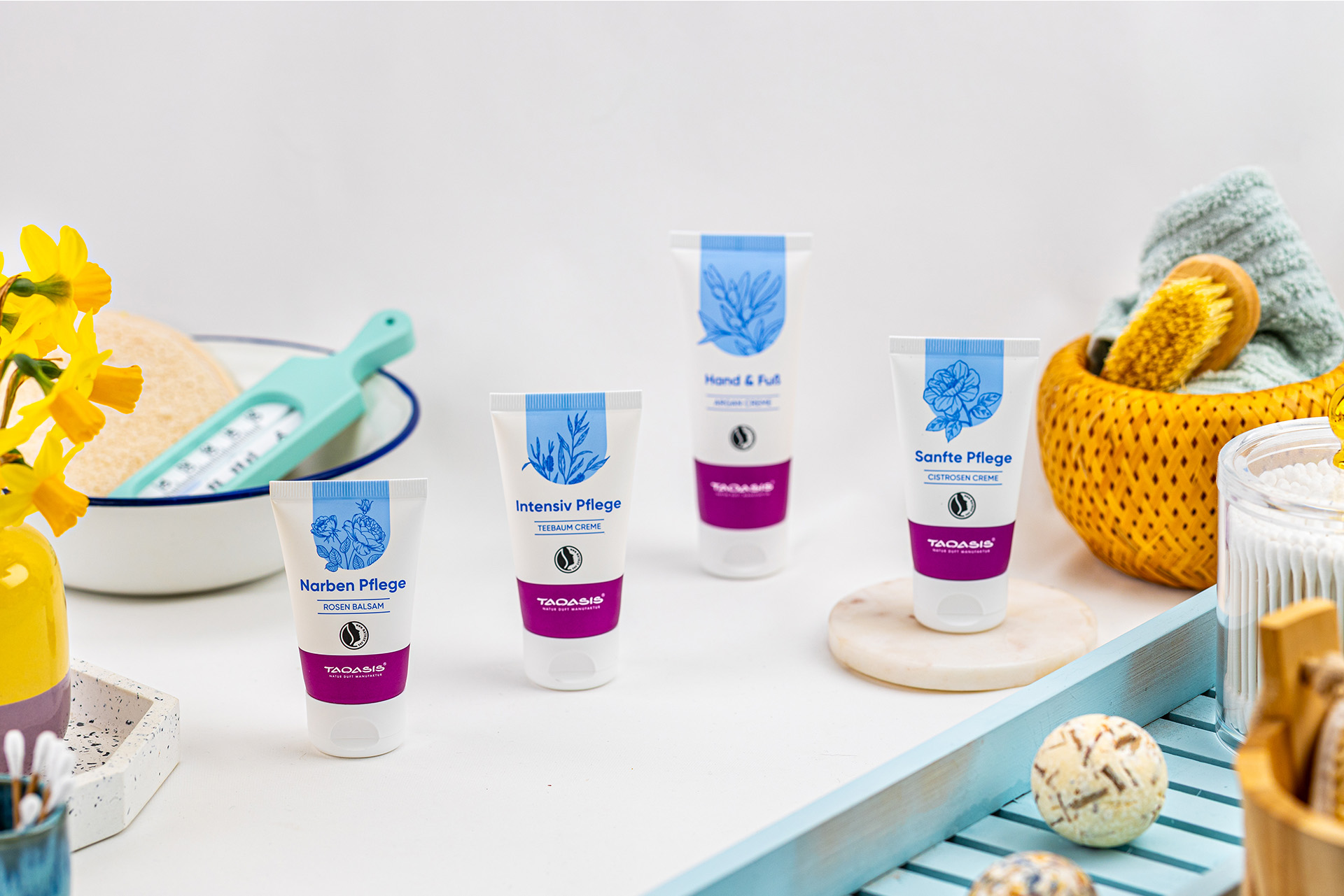 Vier Pflegeprodukte der TAOASIS Hautwohl Serie in einer hellen Badezimmer-Szene mit Blüten, Bürste, Handtuch und weiteren Badeaccessoires.
