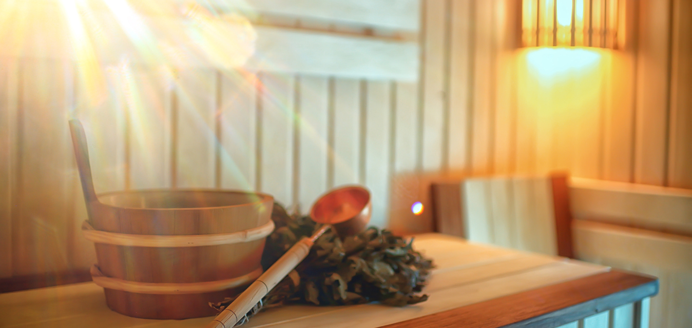 Holzeimer mit Kelle und Birkenzweigen in einer warm beleuchteten Sauna, durchflutet von goldenem Sonnenlicht.