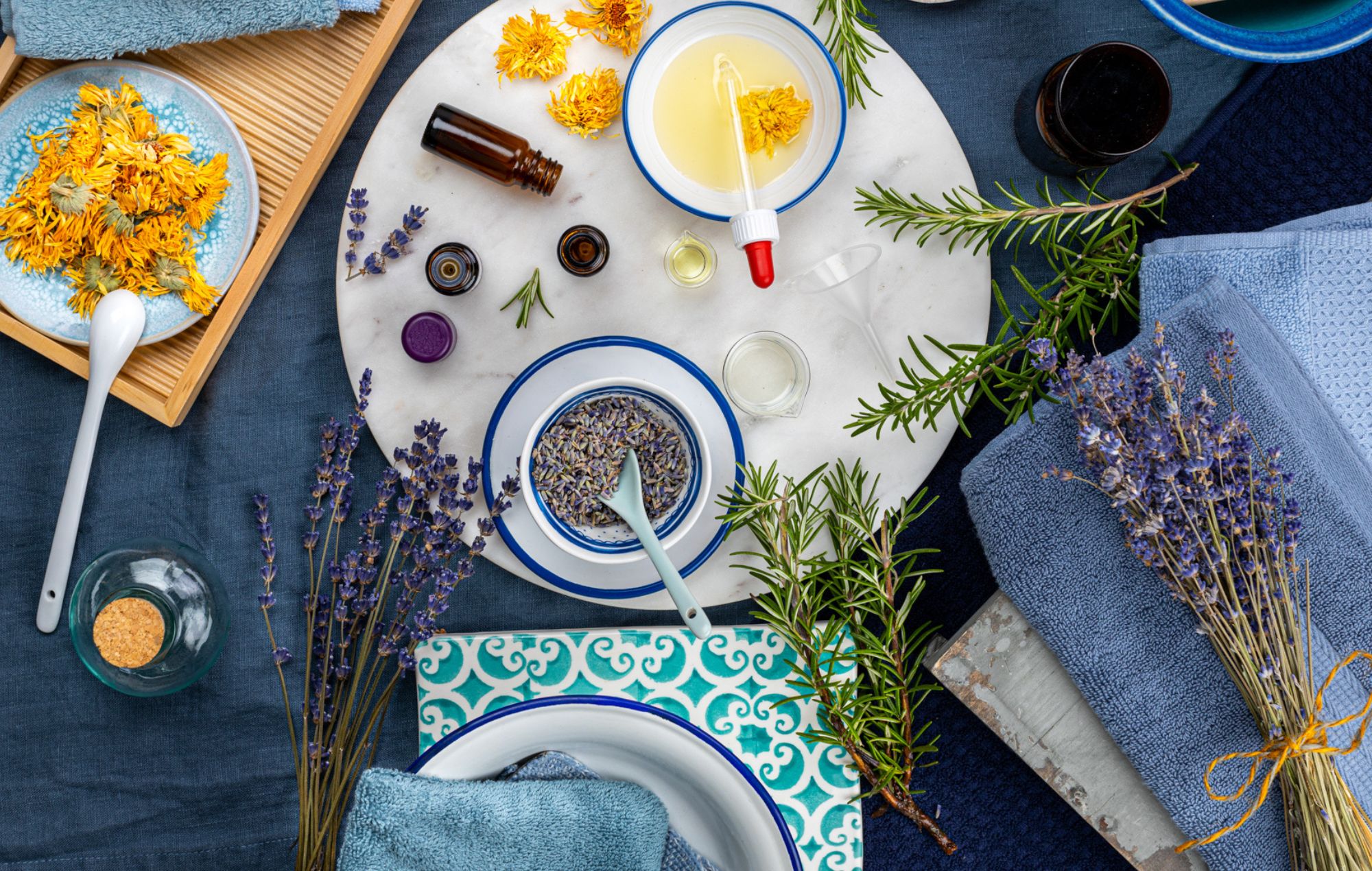 Schalen mit Lavendelblüten und Ringelblumenöl, umgeben von Kräutern, Glasfläschchen und Handtüchern in natürlicher Wellness-Atmosphäre
