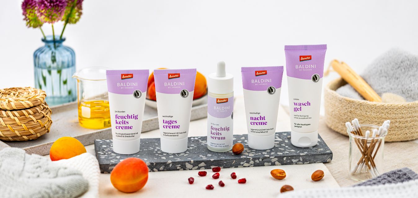 Baldini Naturkosmetik mit Waschgel, Feuchtigkeitsserum, Tagescreme, Feuchtigkeitscreme und Nachtcreme auf Tisch mit natürlichen Zutaten wie Aprikosen und Granatapfelkernen.