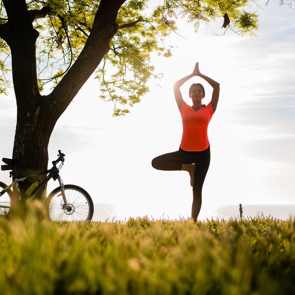 Frau auf einer Wiese, die einen Baum macht. (Yoga) An einem Baum lehnt ein Fahrrad.