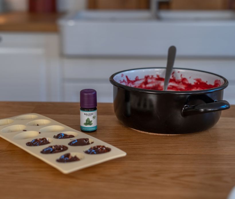 Topf mit Himbeermasse, Pralinenform mit Schokolade und ätherisches Pfefferminzöl auf einem Küchentisch.