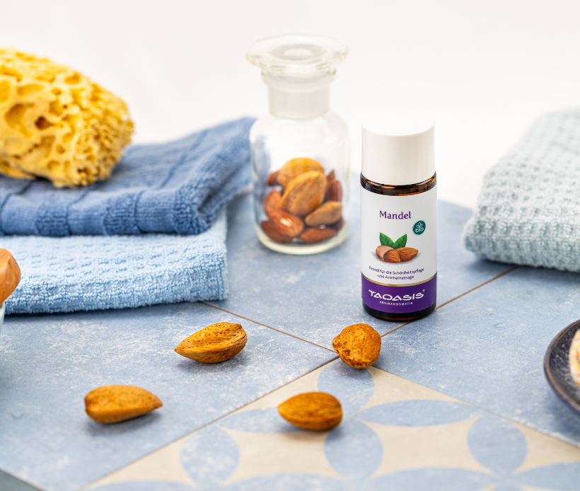 TAOASIS Mandel Basisöl in Glasflasche, umgeben von Mandeln und Wellness-Accessoires.