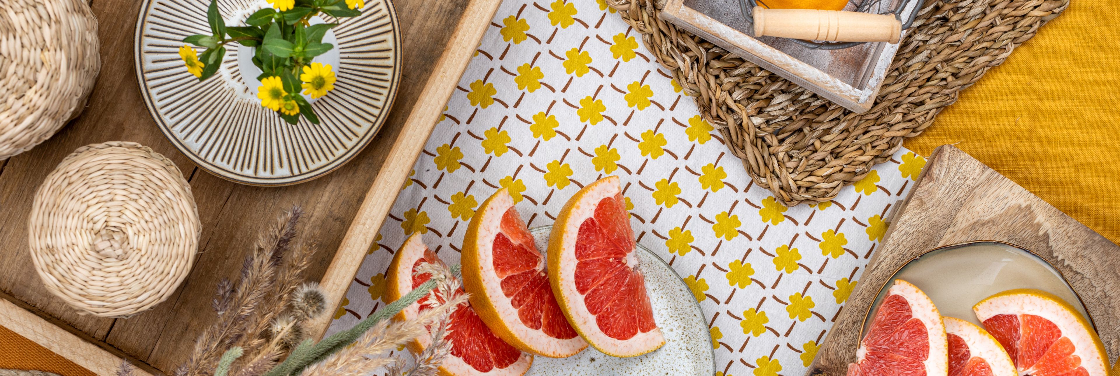 Das Bild zeigt eine sommerlich-warme Szenerie mit frischen Grapefruit-Scheiben, gelben Blüten und natürlichen Materialien wie Holz und Bast – eine harmonische Komposition in Gelb- und Naturtönen, die Frische und Leichtigkeit ausstrahlt.