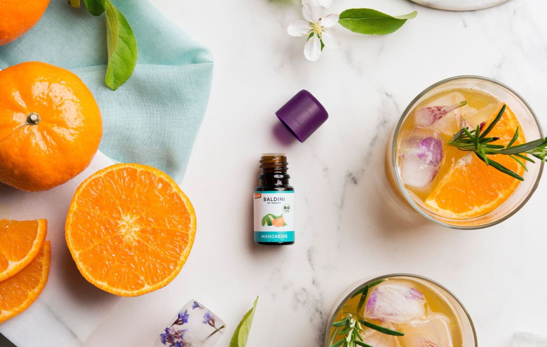 Frische Mandarinen, Bio-Essenz Mandarinenöl und ein Getränk mit Eis, Zitrusfrüchten und Rosmarin dekorativ angerichtet.