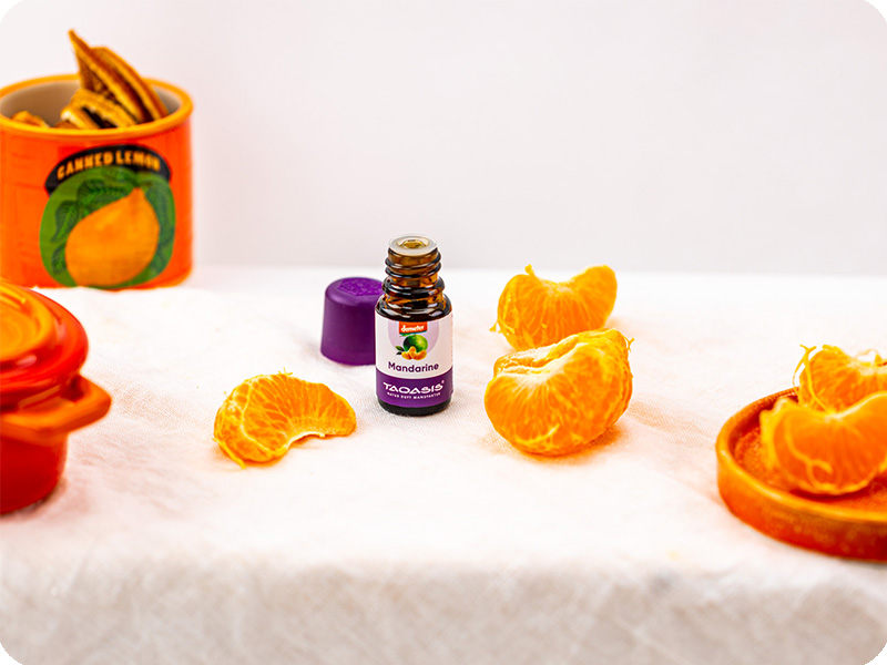 TAOASIS Mandarinenöl mit geschälten Mandarinenstücken und orangefarbenen Accessoires auf hellem Hintergrund.