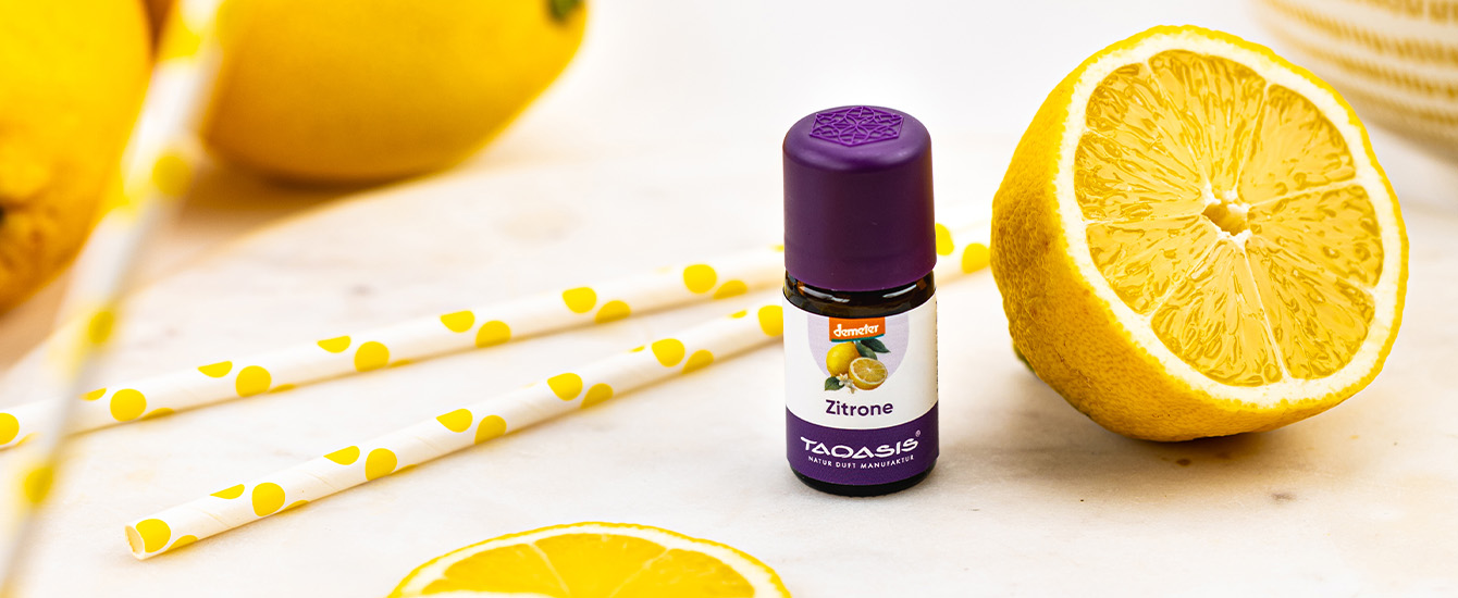 Zitronenöl-Fläschchen mit violettem Deckel neben einer halbierten Zitrone und gelb gemusterten Papierstrohhalmen.