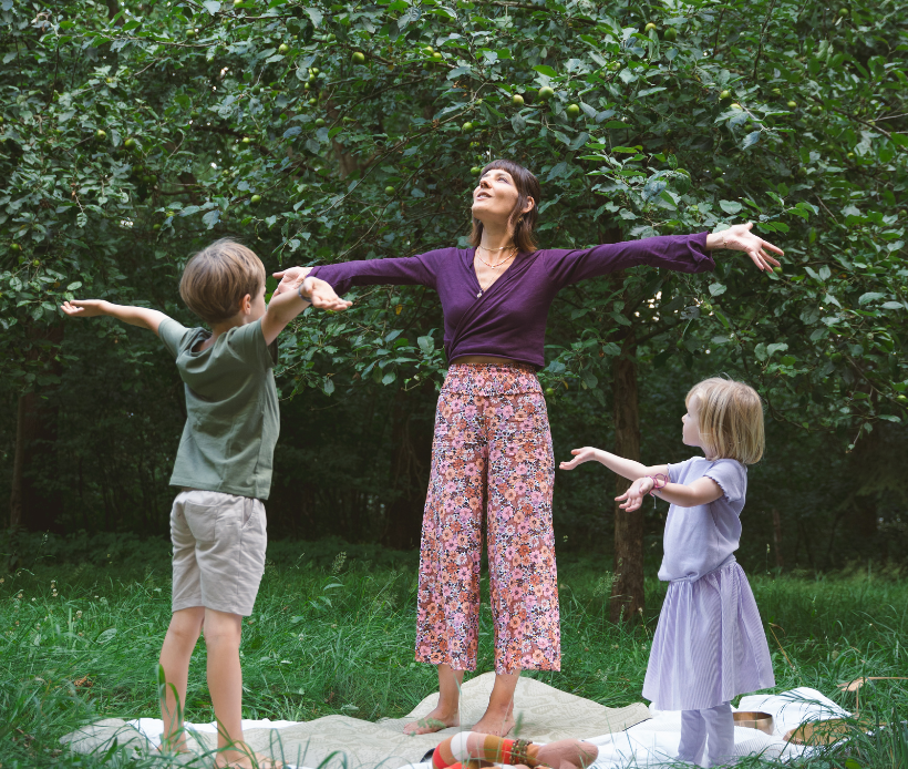 Frau mit zwei Kindern, die Yoga machen. Sie stehen auf einer Decke im Gras vor einem Waldrand. 