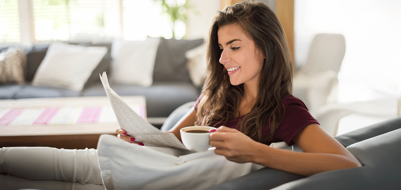 Entspannte Frau sitzt mit einer Tasse in der Hand im Sessel und liest in einer Zeitung.