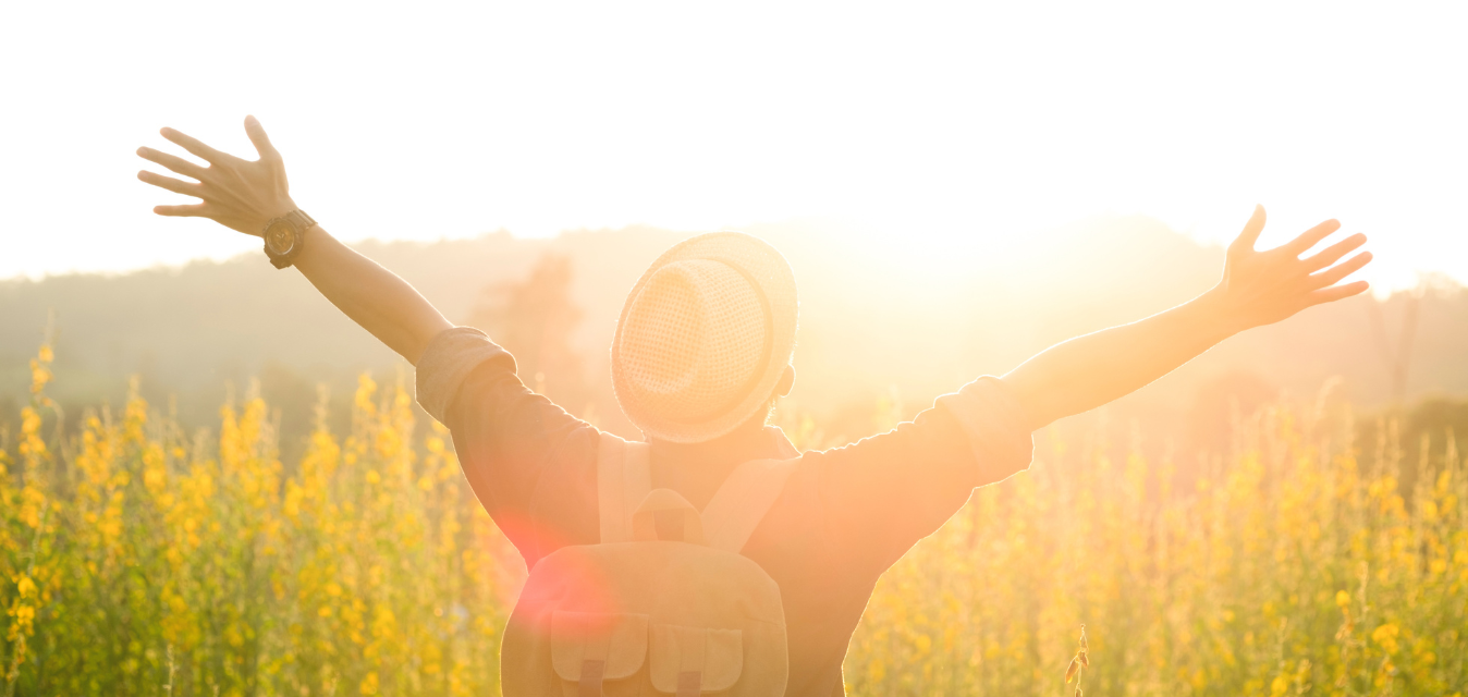 Mann, der einen Hut auf trägt, in der Sonne, im Gras steht und die Arme oben hat. Er trägt einen Rucksack.