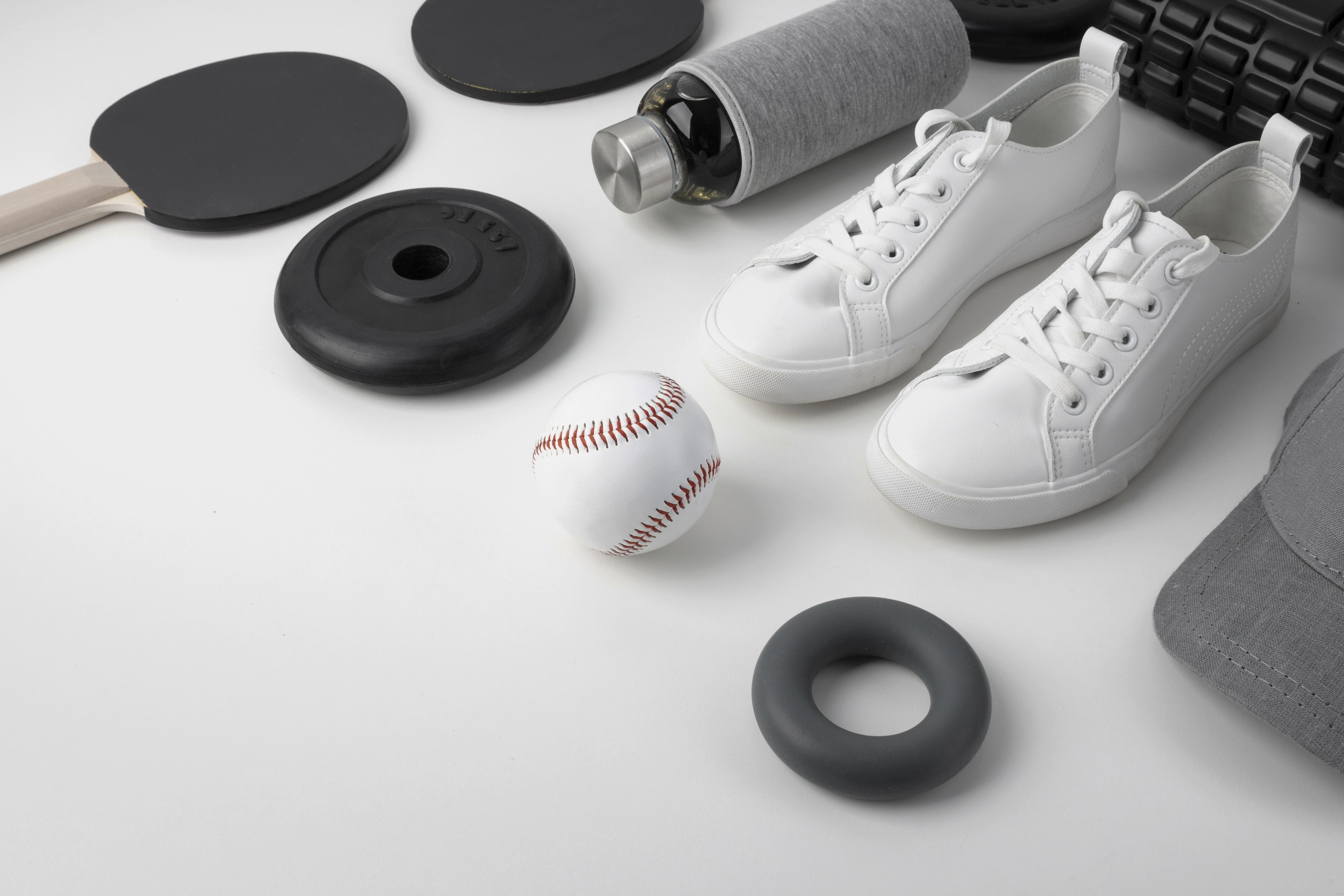 Sportausrüstung auf hellem Hintergrund mit weißen Sneakern, Baseball, Gewichten, Trinkflasche und Fitnesszubehör.