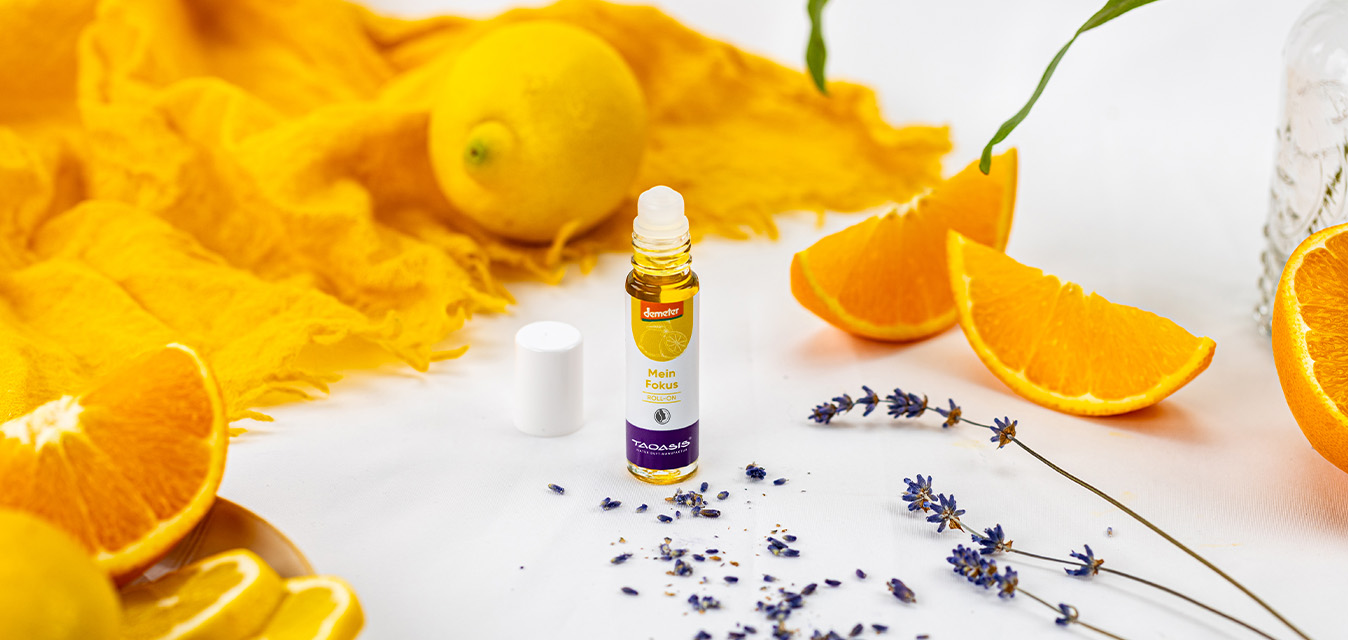TAOASIS Roll-on „Mein Fokus“ umgeben von frischen Orangenspalten, Zitrone, getrocknetem Lavendel und gelbem Stoff.