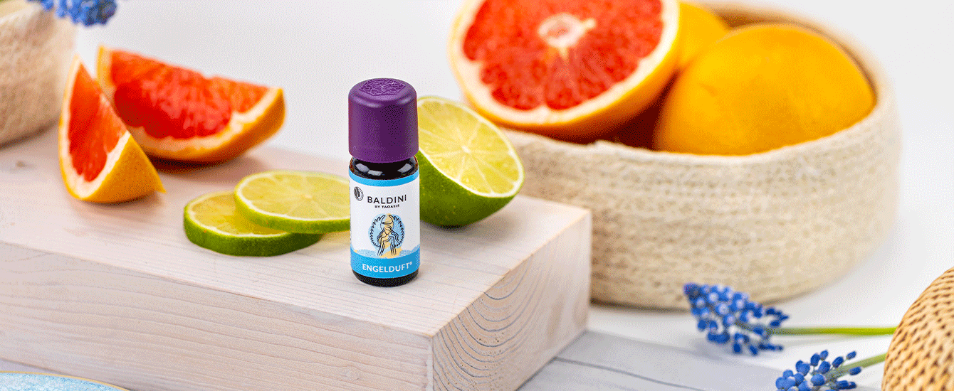 Baldini Engelduft Duftkomposition neben Limetten und Grapefruit auf einem hellen Holzpodest, dekoriert mit blauen Blüten.