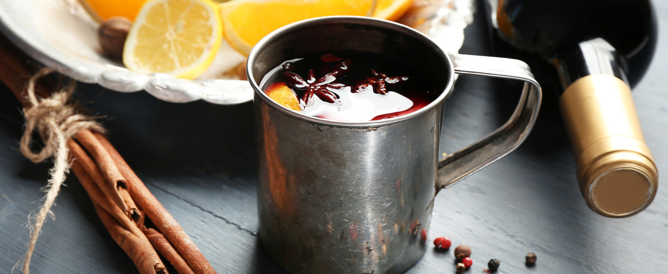 Tasse mit aromatischem Punsch, umgeben von Orangenscheiben, Zimtstangen, Sternanis und winterlichen Zweigen.  Metalltasse mit Glühpunsch, Sternanis und Orangenscheibe, daneben Zimtstangen, Zitrusfrüchte und eine Weinflasche.