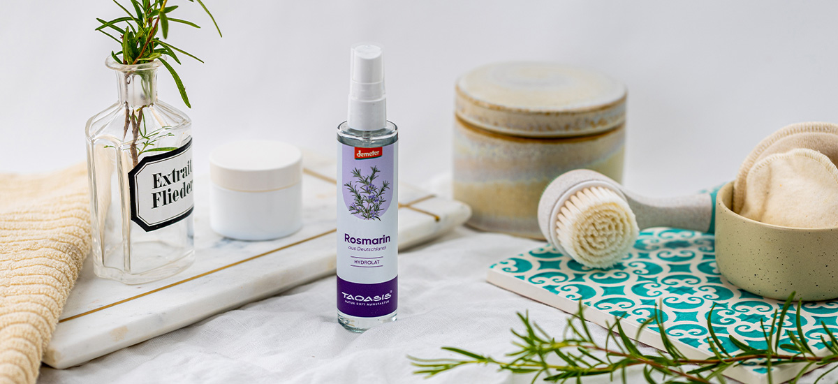 TAOASIS Rosmarin Hydrolat in einer hellen Pflegeszene mit frischem Rosmarin, Gesichtsbürste, Abschminkpads und natürlichen Badaccessoires.