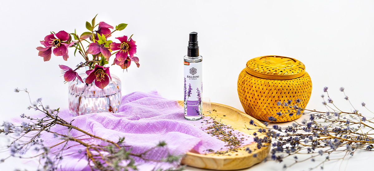 Lavendel-Raumspray von Baldini zwischen violetten Blüten, getrockneten Kräutern, lila Stoff und einem geflochtenen Duftgefäß.