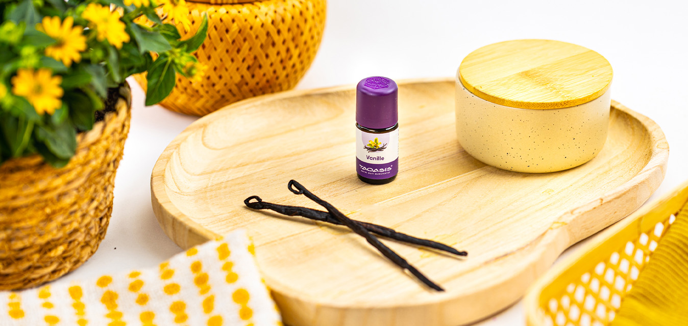Vanilleöl-Fläschchen mit violettem Deckel auf einem Holztablett, dekoriert mit Vanilleschoten und gelben Blüten.