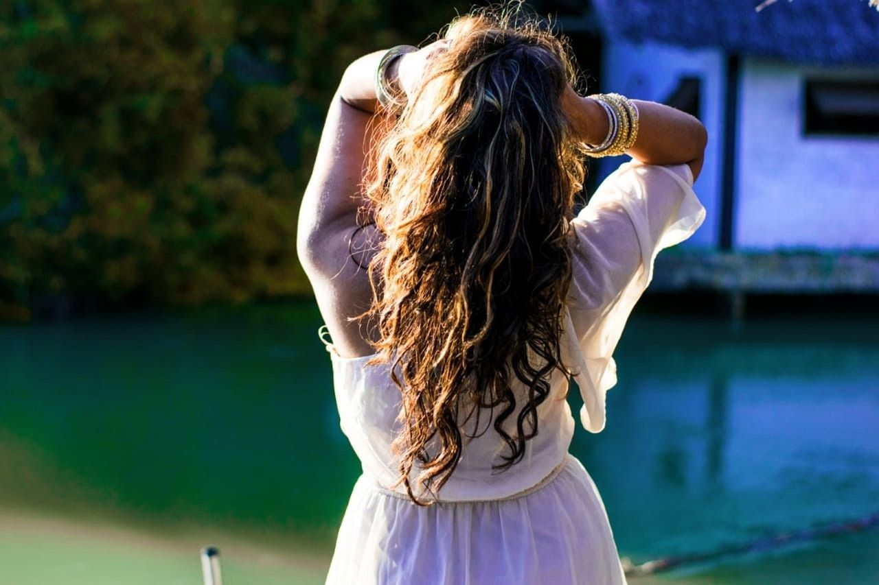 Frau mit langen Haaren steht am Wasser und greift sich in die Haare