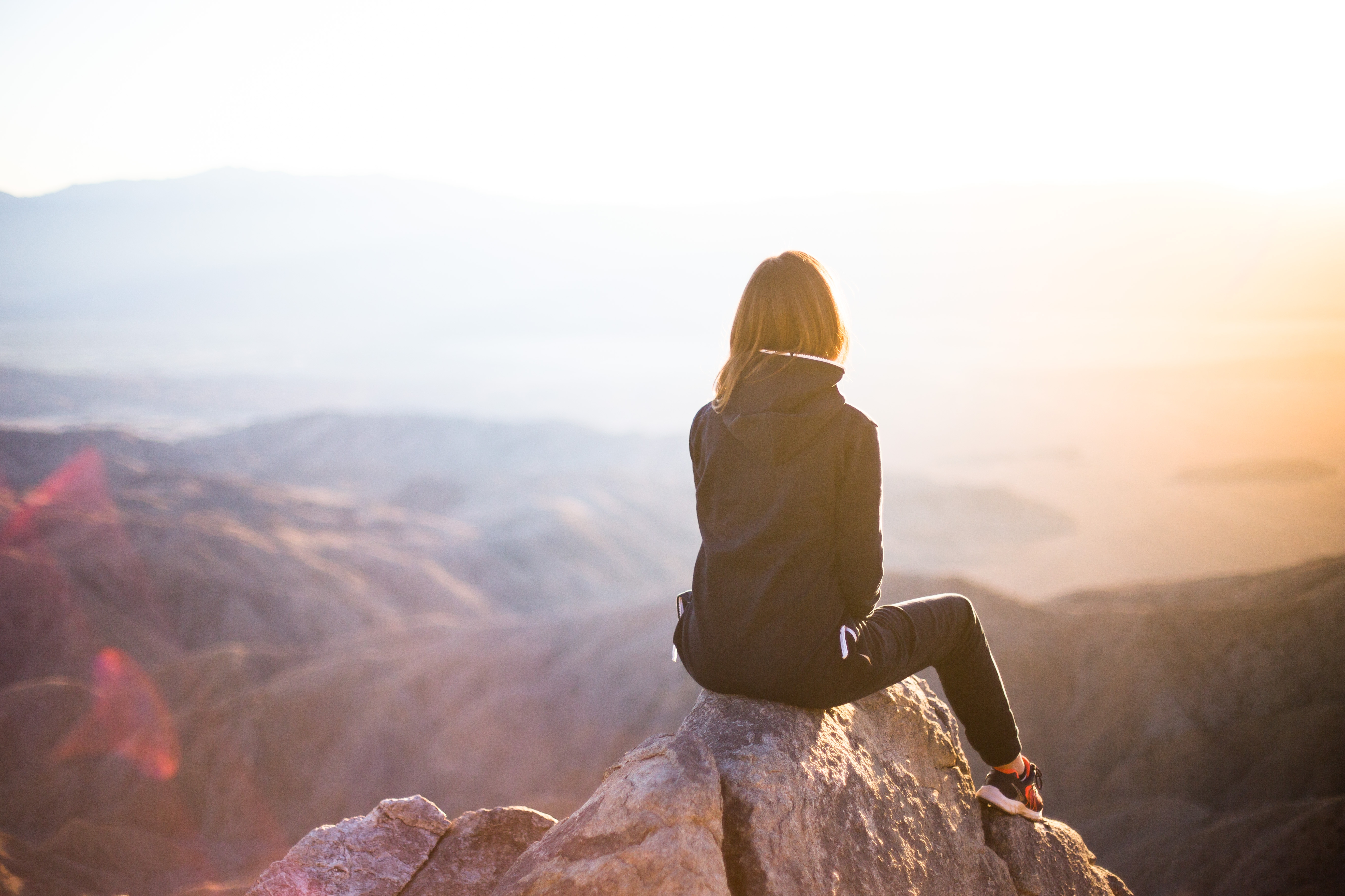 Frau sitzt auf einem Felsen und blickt bei Sonnenaufgang über eine weite Berglandschaft.