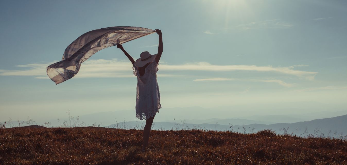 Frau mit Sonnenhut, die auf einer Wiese steht und ein Tuch über ihrem Kopf in den Wind hält. 