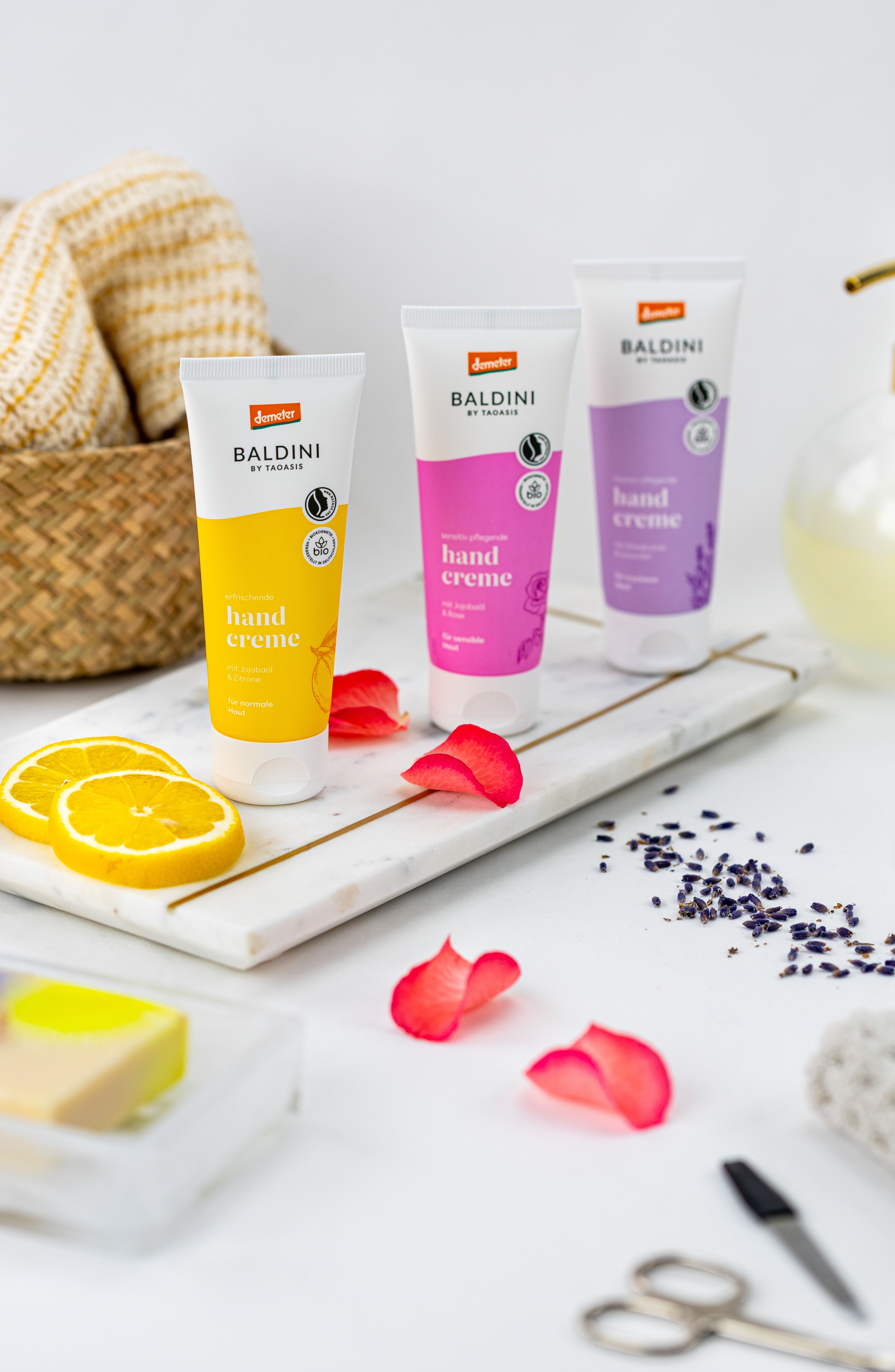 Tablett, darauf stehen 3 Baldini Handcremes (Rose, Zitrus, Lavendel), Blüten liegen auf dem Tisch, sowie eine Kosmetikschere und Pfeile. Im Hintergrund ist ein Korb mit Handtüchern. 