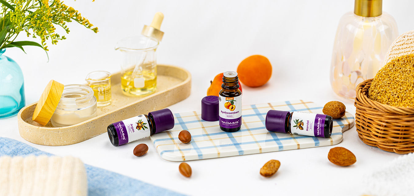 TAOASIS Aprikosenkern-, Argan- und Mandelöl, dekoriert mit Kernen, Pflegeutensilien und natürlichen Accessoires in warmen Gelbtönen.