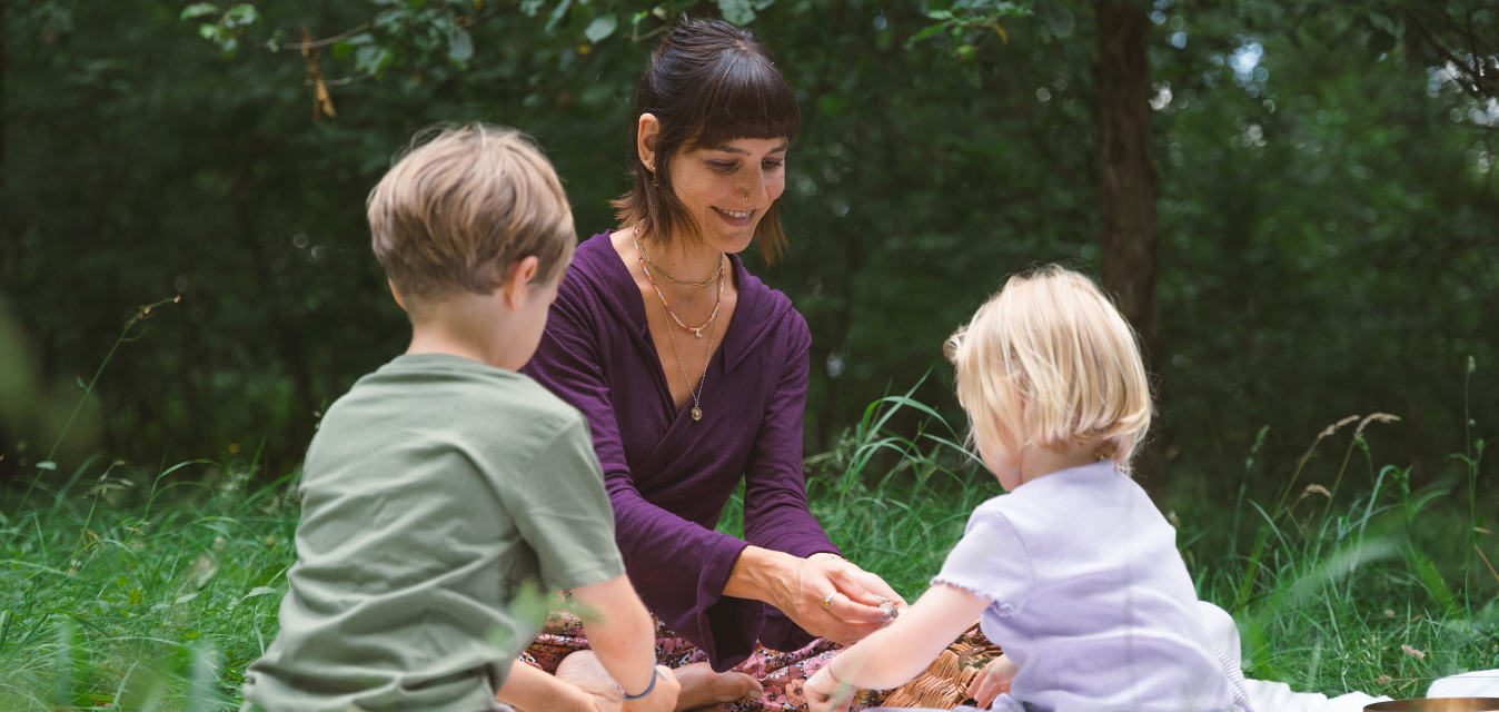 Frau sitzt mir zwei Kinder im Gras. In Hintergrund ist Wald zu erkennen.