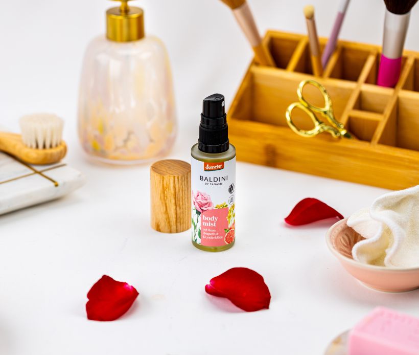 Bodymist Rose umgeben von Rosenblättern und Badezimmerutensilien.