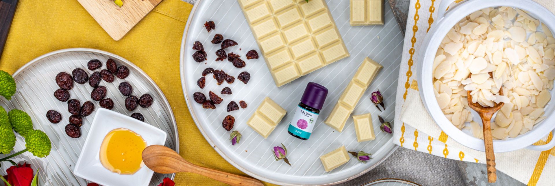 Weiße Schokolade, Mandeln, Rosinen und Rosenöl mit Honig und Blüten dekorativ auf Küchentisch arrangiert.