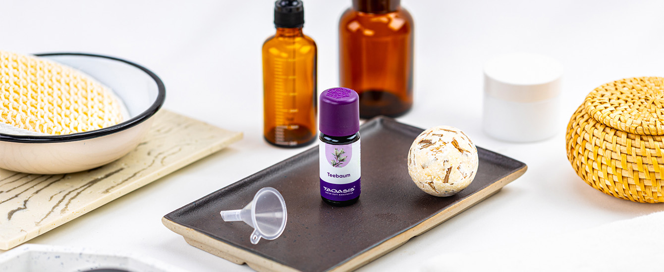 Teebaumöl-Fläschchen mit violettem Deckel auf einer dunklen Keramikschale, daneben ein Badezusatz und Pflegeaccessoires.