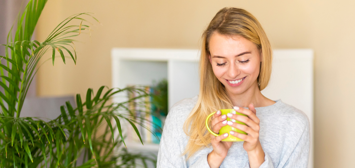 Frau mit Tasse in der Hand. Neben ihr steht eine Pflanze und im Hintergrund ist ein Regal zu sehen.