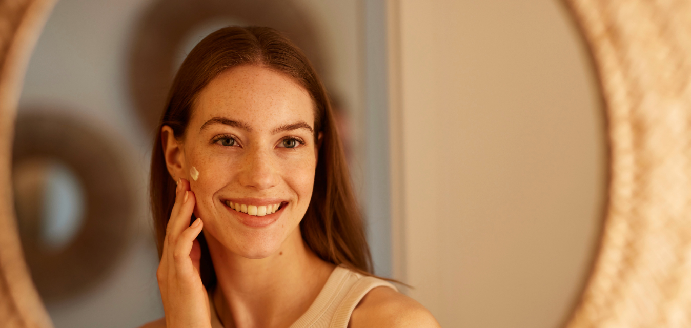 Lächelnde Frau trägt Gesichtscreme auf ihre Wange auf und blickt in einen Spiegel.