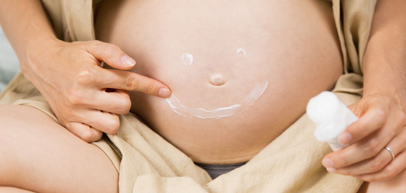Schwangere Frau in beiges Tuch gewickelt. Auf ihrem Bauch ist ein Smiley aus Creme.